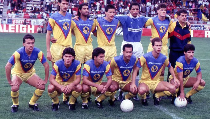 Grupo de once futbolistas masculinos del Club América con uniformes azules y amarillos posando en una cancha verde con un balón de fútbol al frente durante un partido en un estadio con espectadores al fondo y señalización visible del estadio de Raleigh.