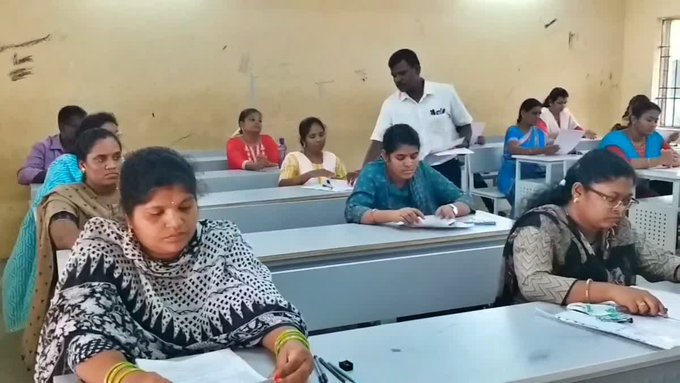 Classroom scene with multiple women and men seated at desks, writing on papers with pens during an examination, wearing traditional and professional attire like sarees and shirts, in a simple yellow-walled room with windows and benches.