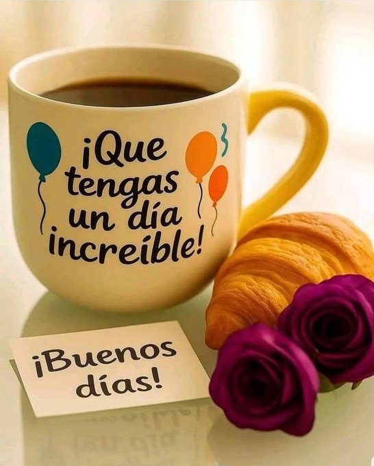 A white ceramic mug with a yellow handle holds dark coffee, featuring printed blue and orange balloons and black text reading Que tengas un dia increible on the side. Next to the mug on a light surface sits a golden croissant and two purple roses with green stems. A small white note with black text Buenos dias is placed nearby.