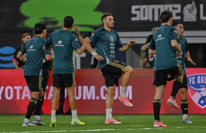 Group of male soccer players wearing green jerseys with sponsor logos and black shorts stand on a green field near a red and white barrier, some raising arms in celebration, others jumping with legs extended, stadium seating and FC Dallas Toyota advertisement visible in background, bright lighting and clear day.