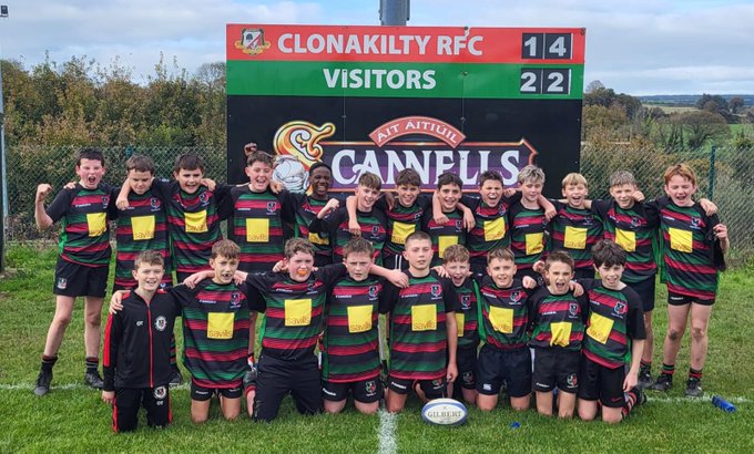 Group of young male rugby players in green and black striped jerseys with yellow accents posing dynamically on a grassy field some kneeling some standing flexing arms and smiling scoreboard in background shows Clonakilty RFC Visitors 14 Highfield RFC 22 rugby ball on ground beside them.