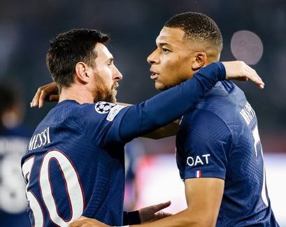 Two soccer players, Lionel Messi and Kylian Mbappé, stand close together on a stadium field at night, wearing matching navy blue Paris Saint-Germain jerseys with the number 10 on Messis back and the UEFA Champions League patch on their sleeves, embracing with arms around each others shoulders while facing each other, blurred stadium lights and crowd in the background.