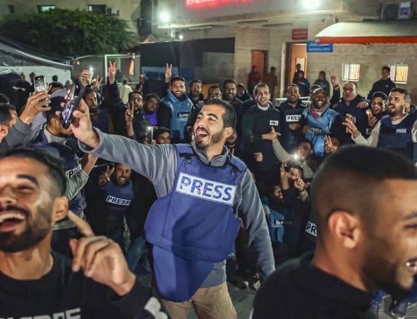 Group of men wearing blue PRESS vests over clothing stand together outdoors at night under lights near buildings and tents with some holding phones and making celebratory gestures with hands raised and smiles on faces in an urban area.