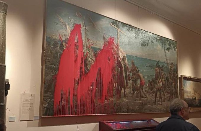 Una gran pintura enmarcada en la pared de un museo representa una escena histórica con figuras vestidas con ropa de época cerca de los barcos y el mar, salpicadas de pintura roja que gotea en el área central. Los elementos circundantes incluyen paredes beige, iluminación tenue, obras de arte adyacentes, una vitrina de madera y un hombre con una chaqueta oscura parado cerca viendo la exhibición.