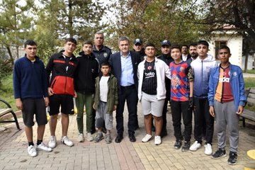 First image shows two men in suits shaking hands outside a shop with tires and boxes nearby, cars parked, and onlookers in background. Second image depicts a man in jacket shaking hands with a suited man near a shop entrance with police officers present and chairs around. Third image features three men in professional attire standing and talking inside a shop near counters and a car visible through door. Fourth image displays a group of about twelve young males and a suited man posing together outdoors on pavement near benches and trees, some wearing soccer jerseys from teams like Barcelona and Fenerbahce.