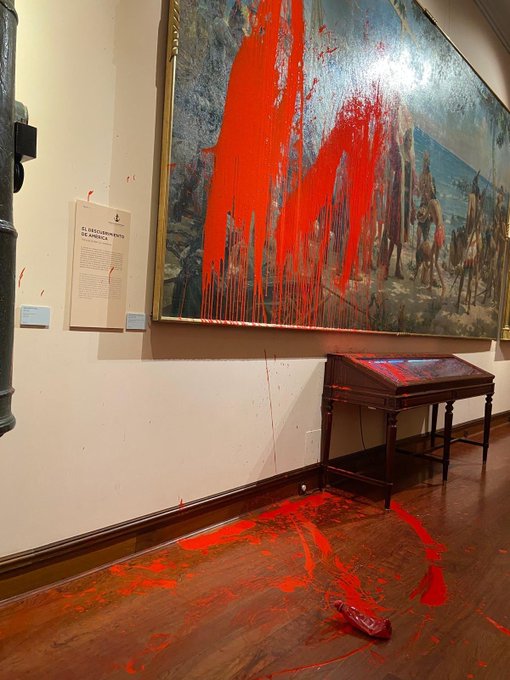 Gran pintura histórica en la pared del museo que representa figuras con atuendos de época con veleros en el fondo, cubiertas de pintura roja biodegradable salpicada que gotea. Rayas de pintura roja en la pared blanca y el piso de madera cercano. Mesa expositora de madera con tapa de cristal en primer plano, parcialmente salpicada de pintura roja. Pequeño cartel en la pared al lado de la pintura con texto. Tubo de metal o barandilla en el lado izquierdo.