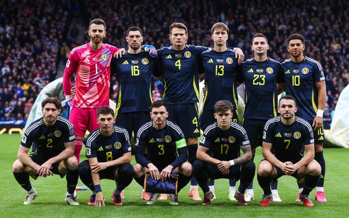 Group of eleven male soccer players from the Scotland National Team posing together on a green grass field in a stadium setting with blurred crowd stands in the background. The players wear matching dark blue jerseys with white numbers including 1, 3, 4, 10, 13, 16, 17, 23, and a goalkeeper in a bright pink jersey numbered 1. They are arranged in two rows, some kneeling and some standing, with arms around shoulders and focused expressions.