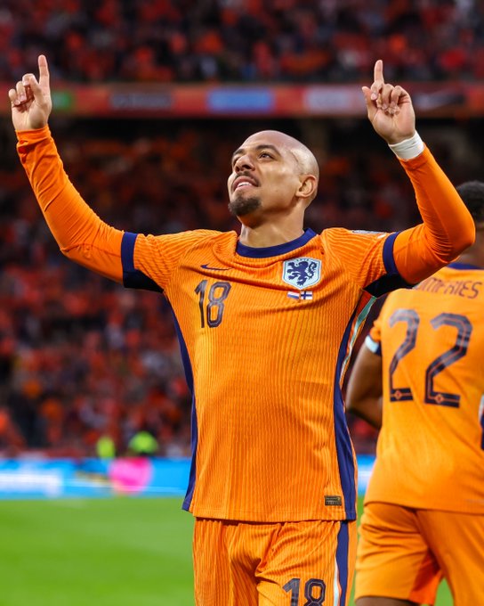 Donyell Malen in orange Netherlands soccer jersey number 18 raises both arms with index fingers up in celebration on green field, another teammate in orange jersey number 22 stands nearby, stadium with blurred crowd and orange banners in background