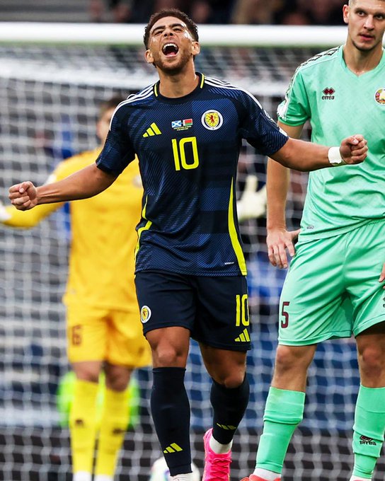 Soccer player Che Adams in dark blue Scotland national team jersey numbered 10 celebrates scoring a goal by raising his arms and shouting in front of the net, with yellow accents on jersey and shorts, pink cleats on feet, another player in green jersey with number 5 stands nearby, stadium seating and goalpost visible in background