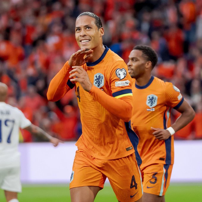 Player in orange Netherlands jersey numbered 4 claps hands smiling on green soccer field, teammate in similar jersey stands nearby with arms crossed, background shows blurred stadium stands filled with orange-shirted fans and another player in white jersey, Dutch lion emblem visible on jerseys.