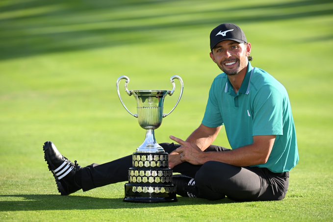 A man wearing a black cap, teal polo shirt, and black pants sits on green grass next to a large silver trophy with gold accents and handles, one hand resting on the base, smiling at the camera, with a golf course in the background.