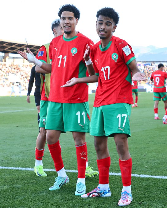 Two young male soccer players from the Morocco national team stand side by side on a green soccer field in a stadium, both with arms extended outward in a celebratory gesture. The player on the left wears a red jersey with green accents numbered 11, green shorts numbered 11, white Puma cleats with colorful designs, and red socks. The player on the right wears a similar red jersey numbered 17, green shorts numbered 17, white Puma cleats, and red socks. Additional teammates in matching red and green uniforms are visible in the background, along with a blurred crowd in the stands.