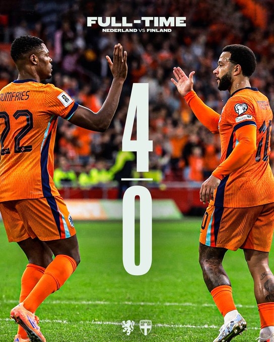 Two male soccer players in orange Netherlands national team uniforms with number 22 on one jersey labeled Dumfries stand on green grass field facing away from camera raising right arms in celebration gesture surrounded by blurred stadium crowd in orange attire full-time scoreboard graphic overlays top showing Netherlands vs Finland 4-0 score lion emblem visible on shorts.