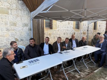 First image shows two men in suits shaking hands outdoors near beige tents with Turkish flags, surrounded by other suited men and autumn trees under a clear sky. Second image depicts four men in formal wear including jackets and glasses shaking hands beside a white tent labeled with Turkish text amid yellow foliage and more tents. Third image features eight older men in suits and jackets seated around white tables under a canopy next to stone walls, some holding phones. Fourth image displays groups of people in dark uniforms seated at tables under a white tent with Turkish text, near purple trees and benches in a park-like setting.