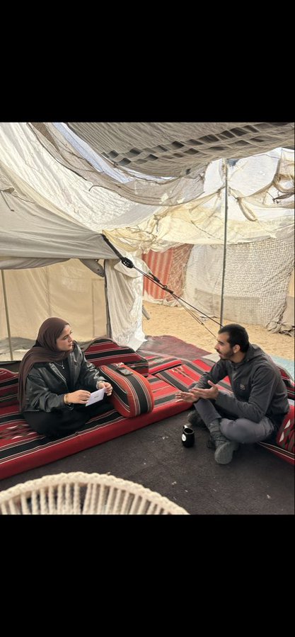 A woman wearing a brown hijab and black clothing sits on a red and white striped mattress inside a tent made of white fabric supported by poles, with a metal container nearby. A man in a gray hoodie and pants sits opposite her on the same mattress, holding a phone. The ground is sandy, and additional tent structures and a white chair are visible in the background.