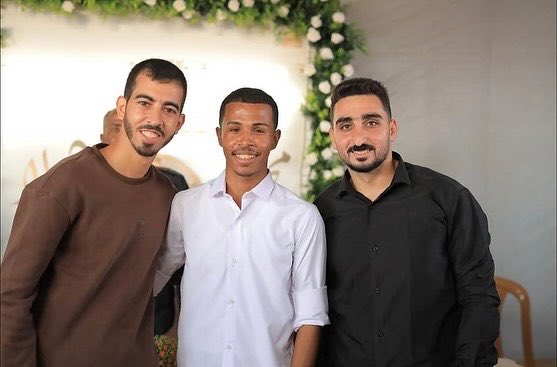 Three young men of Middle Eastern descent stand closely together in a group pose all smiling warmly at the camera. The man on the left wears a brown short-sleeved shirt has short dark hair and a trimmed beard. The middle man is dressed in a white short-sleeved button-up shirt has close-cropped hair and a friendly expression. The man on the right sports a black short-sleeved shirt has curly dark hair and a mustache. Behind them is a decorative backdrop of green leaves and white roses suggesting an event setting.