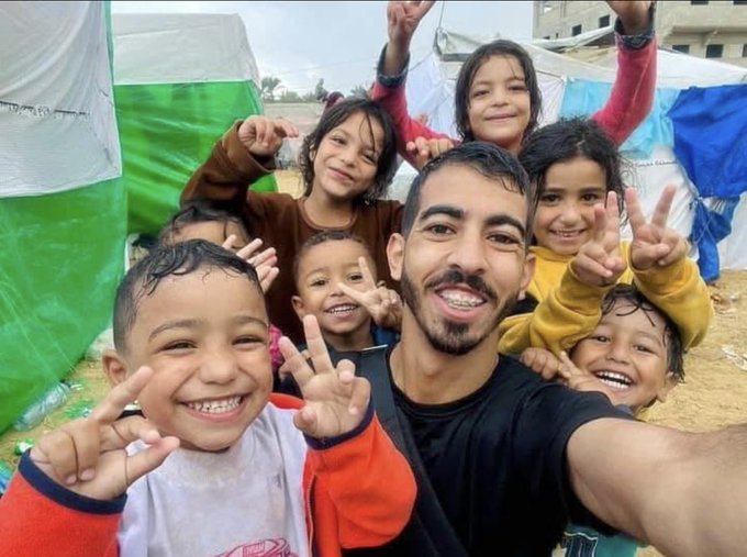 A group photo shows a man with a beard and short hair smiling while holding a young child on his shoulders, surrounded by several smiling children of various ages making peace signs with their fingers. The children wear colorful casual clothes like hoodies and t-shirts. They stand in front of white tents with green and blue fabric flaps in a sandy outdoor area under a partly cloudy sky.