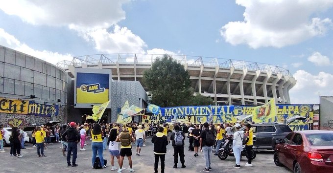 Exterior del estadio con grandes pancartas amarillas y azules que dicen decoraciones de estilo AZTECA y LA MONUMENTAL GRAFITI en las paredes, multitudes de personas, algunas con camisetas amarillas, de pie alrededor de autos estacionados, incluido un SUV rojo y motocicletas bajo un cielo azul parcialmente nublado, árboles visibles cerca