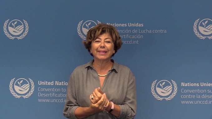 A woman with curly brown hair smiles while standing in front of a blue backdrop featuring repeated United Nations logos and text in French and Spanish reading Nations Unies and Naciones Unidas along with Convention des Nations Unies contre la desertification and www.unccd.int.