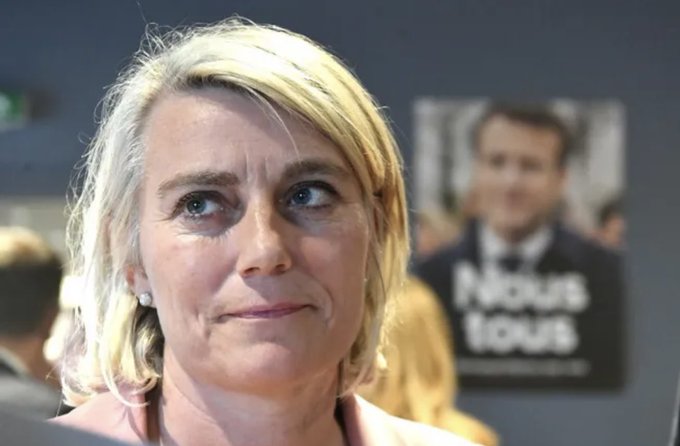 A woman with blonde hair wearing earrings and a pink top stands in a room with a blurred background featuring a poster of a man in a suit and a sign reading NOUS TOUS in blue and white letters alongside other indistinct figures