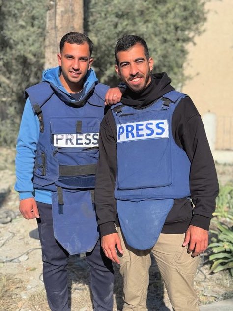 Two men stand side by side outdoors near trees and plants, both wearing blue hoodies under black tactical vests labeled PRESS in white letters, with one man draping an arm over the others shoulder, both in pants and appearing in a professional press context.