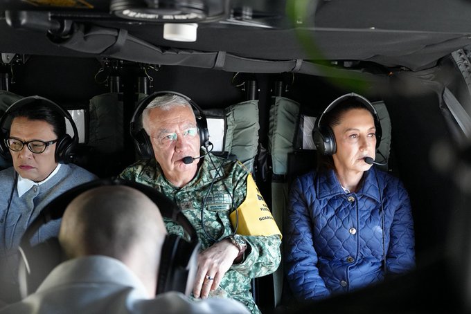 Dentro de la cabina de un helicóptero, cuatro personas usan auriculares y se sientan en asientos. Una mujer con anteojos y ropa gris está a la izquierda. Junto a ella, un hombre mayor con uniforme militar y un brazalete amarillo habla por su micrófono. Otra mujer con una chaqueta azul con un micrófono está a la derecha. Un hombre a la vista parcial se sienta en primer plano. El equipo militar y el equipo son visibles a su alrededor.