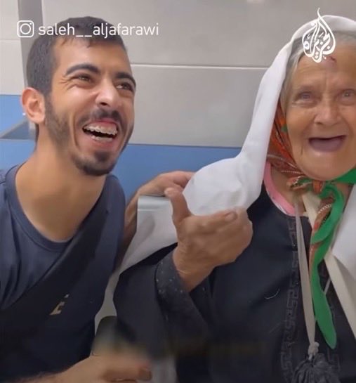 Young man with short dark hair and beard wearing a dark t-shirt smiles widely while hugging an elderly woman from the side. The woman has white hair covered by a white headscarf and wears a colorful scarf around her neck, smiling broadly. They sit on blue seats against a white tiled wall. Al Jazeera logo visible on clothing. Instagram watermark present at bottom.