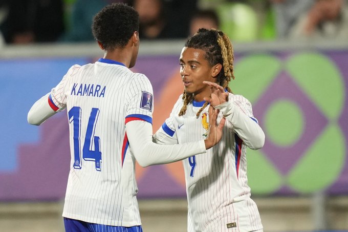 Dos jugadores de fútbol franceses con uniformes blancos con detalles azules y números 14 y 9 celebran chocando los cinco en un campo del estadio rodeado de una multitud borrosa y un campo verde. Un jugador tiene rastas y apunta hacia adelante mientras que el otro sonríe y gesticula con el brazo levantado. El fondo muestra los asientos y las pancartas del estadio.