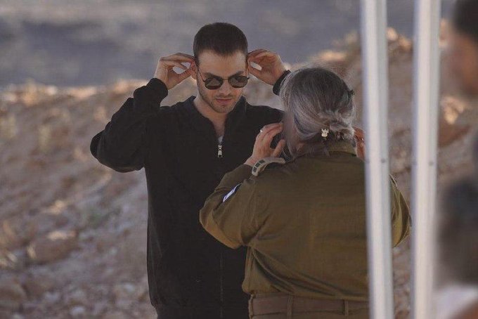 First image shows a young man in black zip-up jacket and sunglasses adjusting his eyewear while a woman with gray hair in ponytail wearing green military uniform stands facing him in a dusty outdoor area with debris and a metal pole behind. Second image captures the same young man in black jacket raising his hand in greeting toward the woman in green uniform as two blurred men in dark clothing and one in green uniform with Israeli flag patch stand nearby under a blue tent in a rugged terrain with rocks and equipment.