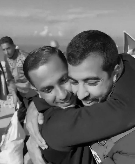 Black-and-white photo shows two men in military uniforms embracing closely outdoors with tents and equipment in the background under a clear sky. Another man stands nearby observing. The scene conveys camaraderie and emotion.