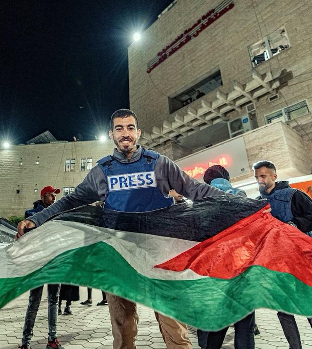 A man in a blue press vest and gray hoodie smiles while holding a large Palestinian flag featuring green white black and red stripes in front of a beige building at night under bright lights. Other individuals in similar vests and red caps stand nearby in an urban setting with architectural elements visible.