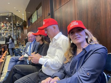 First image displays a red baseball cap with white text reading Trump the peace president held in a hand against a blurred background of a modern legislative chamber featuring wooden paneling، chairs، and screens. Second image captures a diverse group of attendees in formal attire seated in rows within the chamber، several wearing red Trump-style caps، including individuals in suits and one in a leopard-print outfit، with architectural elements like railings and lighting visible. Third image shows four people seated closely، two men and two women in business casual clothing، with the men wearing red Trump the peace president caps، one woman smiling and wearing earrings، set against wooden walls and seating areas in the chamber.