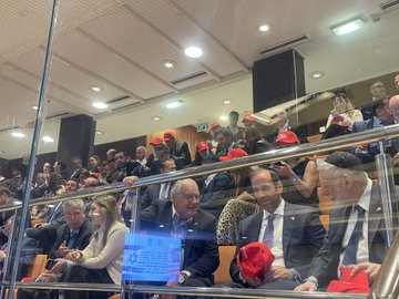 First image displays a red baseball cap with white text reading Trump the peace president held in a hand against a blurred background of a modern legislative chamber featuring wooden paneling، chairs، and screens. Second image captures a diverse group of attendees in formal attire seated in rows within the chamber، several wearing red Trump-style caps، including individuals in suits and one in a leopard-print outfit، with architectural elements like railings and lighting visible. Third image shows four people seated closely، two men and two women in business casual clothing، with the men wearing red Trump the peace president caps، one woman smiling and wearing earrings، set against wooden walls and seating areas in the chamber.