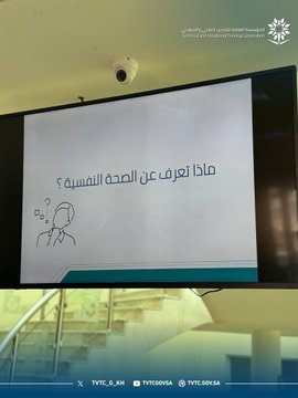 First image shows a table with multiple blue water bottles, white cups, and snacks on a white cloth in a modern room with geometric wall patterns and a stand holding a framed QR code. Second image displays a screen presenting a question about mental health in Arabic with illustrative icons and a cartoon figure, set in an indoor space with a surveillance camera and railing. Third image features a wall with the TVTC logo and Arabic-English text for Technical and Vocational Training Corporation, alongside a table of water bottles and a stand with a QR code. Fourth image depicts several bouquets of white roses wrapped in green paper with Arabic text on ribbons symbolizing awareness.