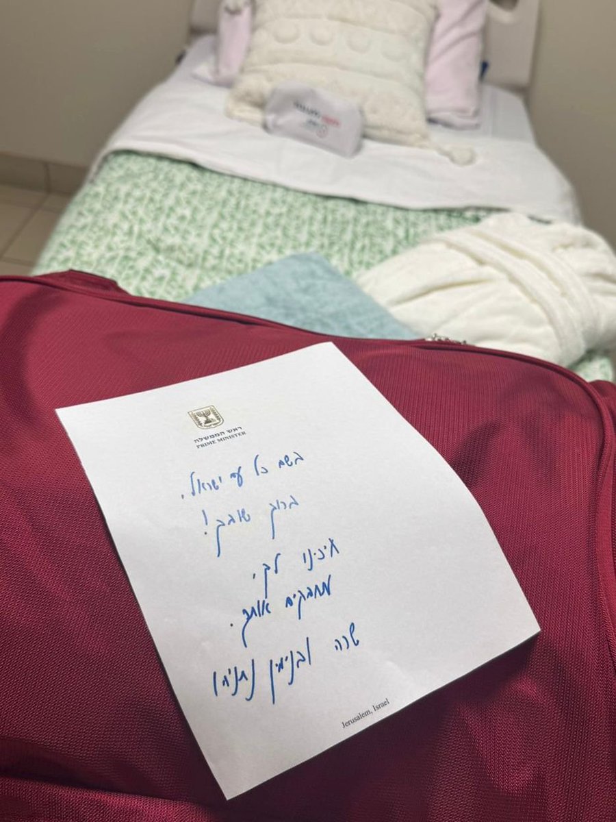 A red suitcase lies open on a green bedspread in a bedroom with white walls and soft lighting. A white paper note with handwritten Hebrew text in blue ink rests on the suitcase, partially visible. Nearby are white and pink pillows on the bed, and a white piece of furniture in the background.