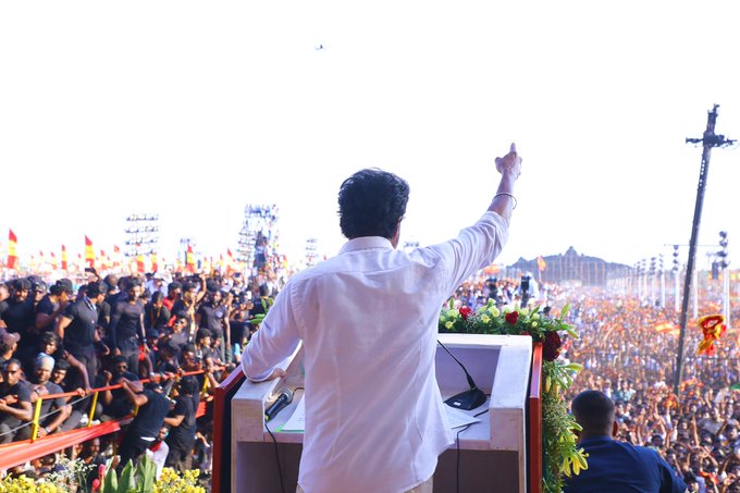 A man in white shirt stands on a large outdoor stage raising his right arm while speaking into a microphone with a crowd of people gathered below holding orange flags and banners under a clear sky with decorative elements like flowers and poles visible in the background