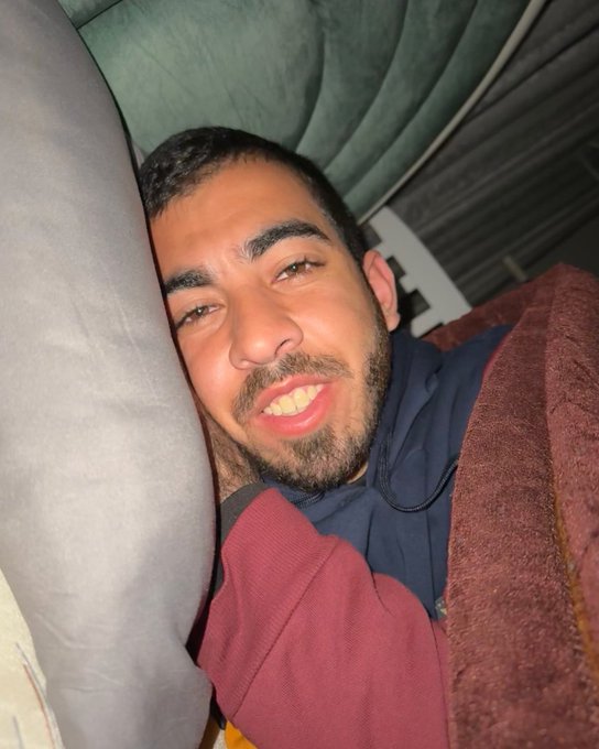 A young man with short dark hair, a beard, and a slight smile rests his head on a white pillow in a dimly lit indoor setting. He wears a dark hoodie and has earbuds around his neck. The background includes a green cushioned surface and maroon fabric, suggesting a bed or couch area.