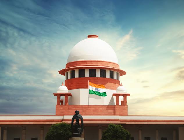 Red sandstone building with white dome and arches under blue sky with Indian flag flying. Statues on the roof and green shrubs in front.