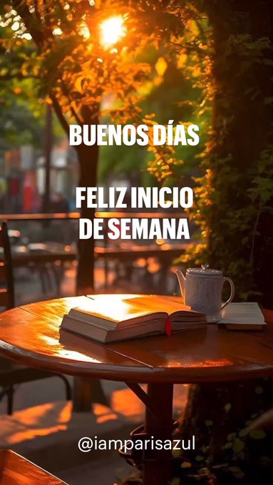 A round wooden table sits outdoors under green trees with sunlight filtering through leaves creating a warm glow. An open book lies on the table with pages spread. A white teapot rests beside the book. Text overlay reads BUENOS DÍAS and FELIZ INICIO DE SEMANA in large letters. Empty chairs surround the table in a park-like setting.