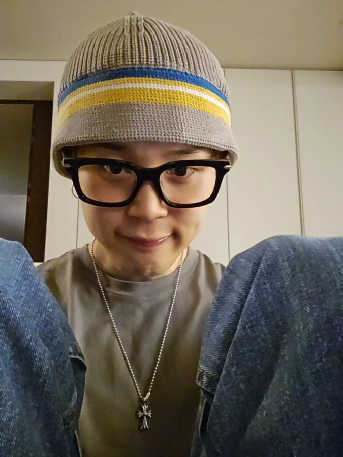 A young Asian individual with short dark hair sits indoors against a white wall, wearing a gray knit beanie with horizontal yellow and blue stripes, black rectangular glasses, a loose light gray t-shirt, a silver chain necklace featuring a small cross pendant, and relaxed blue jeans with cuffs rolled up, smiling softly with hands visible in the foreground.