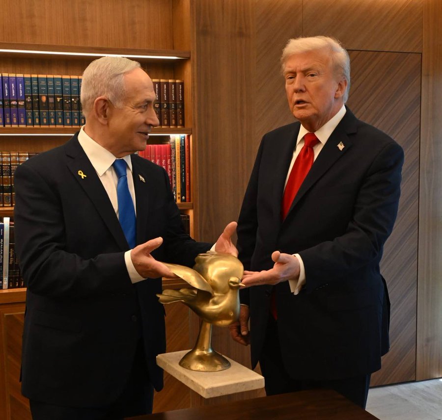 身穿深色西装、打着蓝色领带的本杰明·内塔尼亚胡（Benjamin Netanyahu）在底座上拿着一个金色的鸽子雕塑，将其伸向身穿深色西装、打着红色领带的唐纳德·特朗普，两人都站在一个木板房间里，房间里有书架，后面有一张木桌。