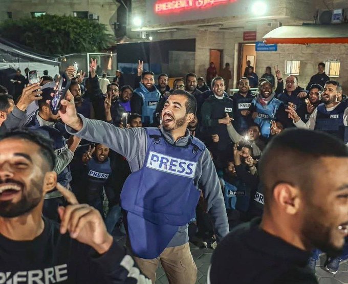 Group of men wearing blue press vests and dark security uniforms stand outdoors at night in front of a beige building with an illuminated emergency room sign above the entrance. Several men raise their hands in celebration with smiles on their faces while holding phones and bottles. Palm trees and tents are visible in the background under bright overhead lights.
