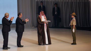 First image shows a large stage with a screen displaying Expo 2025 Closing Ceremony in English and Japanese, featuring five men in suits and one in traditional Saudi thobe standing together under curved architecture. Second image depicts four young men in khaki uniforms and hats holding and presenting a white flag with a blue circle emblem on a stage with golden poles and audience below. Third image captures two men in suits and one in Saudi thobe and red checkered headscarf shaking hands, with a boy scout in uniform standing nearby, against a backdrop of curtains and screens. Fourth image shows elderly men in suits clapping, a man in thobe holding a document, and a boy scout saluting, positioned on a wide stage with draped backgrounds.