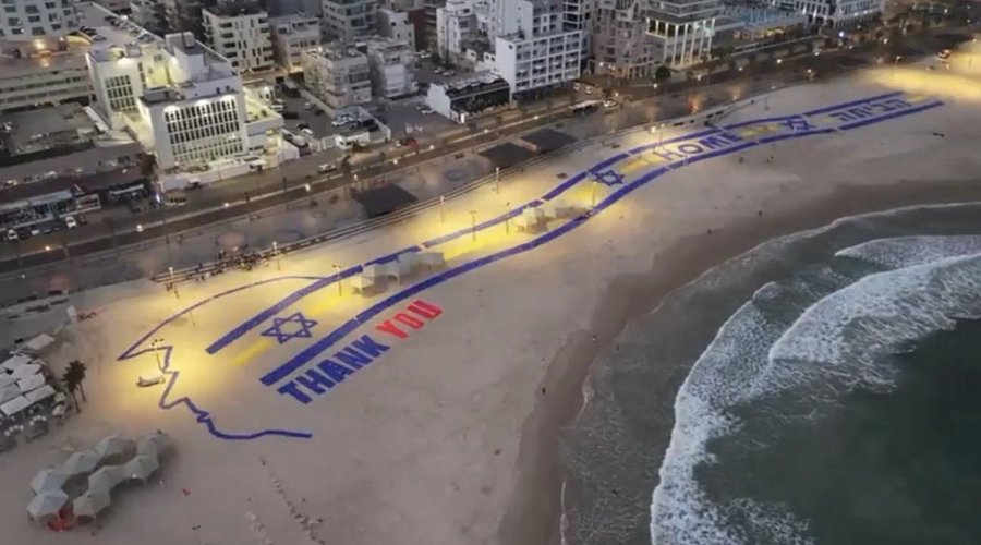 Aerial perspective over Tel Aviv beach at night shows buildings and shoreline with ocean waves. Large illuminated display on sand features blue and white Israeli flag stripes forming text Thank you in red and blue letters alongside a profile outline resembling Donald Trump. Surrounding area includes parked vehicles and tents near the water edge.
