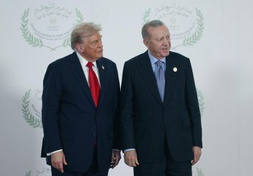 First image shows two men in dark suits standing side by side with thumbs up gestures, one wearing a red tie and American flag pin, the other a blue tie and small badge, against a white backdrop with Arabic script and laurel designs. Second image depicts the same men shaking hands formally. Third image captures them facing each other closely with serious expressions. Fourth image shows them standing together smiling slightly. The backdrop features symmetric Arabic text and olive branch motifs.