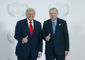 First image shows two men in dark suits standing side by side with thumbs up gestures, one wearing a red tie and American flag pin, the other a blue tie and small badge, against a white backdrop with Arabic script and laurel designs. Second image depicts the same men shaking hands formally. Third image captures them facing each other closely with serious expressions. Fourth image shows them standing together smiling slightly. The backdrop features symmetric Arabic text and olive branch motifs.