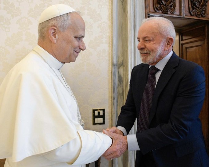 Pope Francis wearing white cassock and zucchetto stands in an ornate interior with beige walls and wooden doors shaking hands with Lula da Silva in dark suit red tie and white shirt both elderly men with white hair smile facing each other a wall-mounted electrical outlet is visible nearby