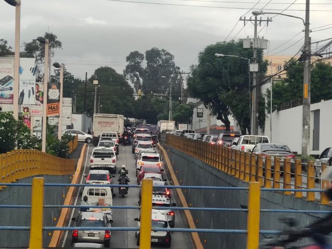 A wide elevated viaduct road in an urban area shows heavy traffic congestion with numerous cars, trucks, and motorcycles stopped or slowly moving in both directions. Yellow guardrails line the sides, with trees, billboards, and buildings visible along the roadside. Overcast sky and utility poles are present, indicating a busy city thoroughfare toward Charcas in zone 11.