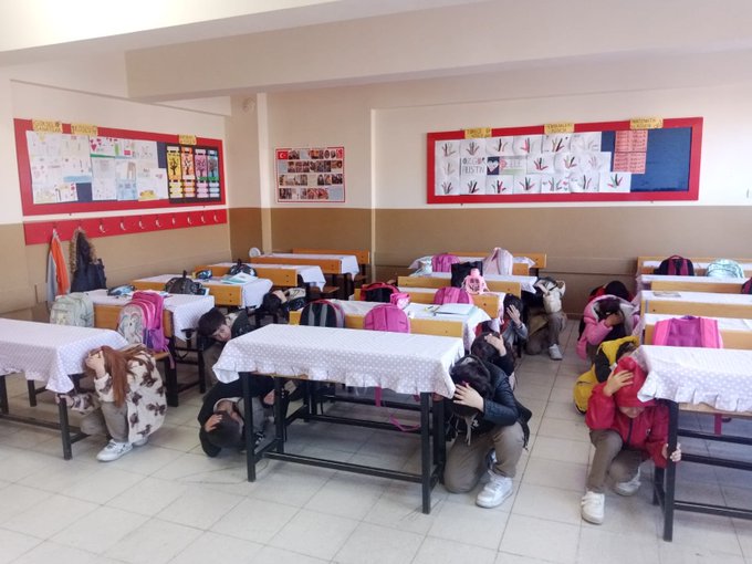 First image shows a classroom with young girls in colorful uniforms and headscarves sitting around a table covered with a white cloth some wearing animal-themed costumes like cow patterns others with backpacks on chairs bulletin boards with drawings and educational posters on walls red-trimmed displays and hanging clothes. Second image depicts students mostly boys and girls in blue uniforms kneeling or sitting on the floor in a classroom with desks pushed aside heads covered with arms or cloths simulating hiding during an earthquake gray board with posters water bottles and backpacks scattered around. Third image captures multiple students in similar uniforms crouching under or beside desks in a spacious classroom tiled floor gray board with decorations backpacks and water bottles on desks some children covering heads with arms or cloths during the drill exercise.