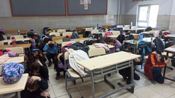 First image shows a classroom with young girls in colorful uniforms and headscarves sitting around a table covered with a white cloth some wearing animal-themed costumes like cow patterns others with backpacks on chairs bulletin boards with drawings and educational posters on walls red-trimmed displays and hanging clothes. Second image depicts students mostly boys and girls in blue uniforms kneeling or sitting on the floor in a classroom with desks pushed aside heads covered with arms or cloths simulating hiding during an earthquake gray board with posters water bottles and backpacks scattered around. Third image captures multiple students in similar uniforms crouching under or beside desks in a spacious classroom tiled floor gray board with decorations backpacks and water bottles on desks some children covering heads with arms or cloths during the drill exercise.